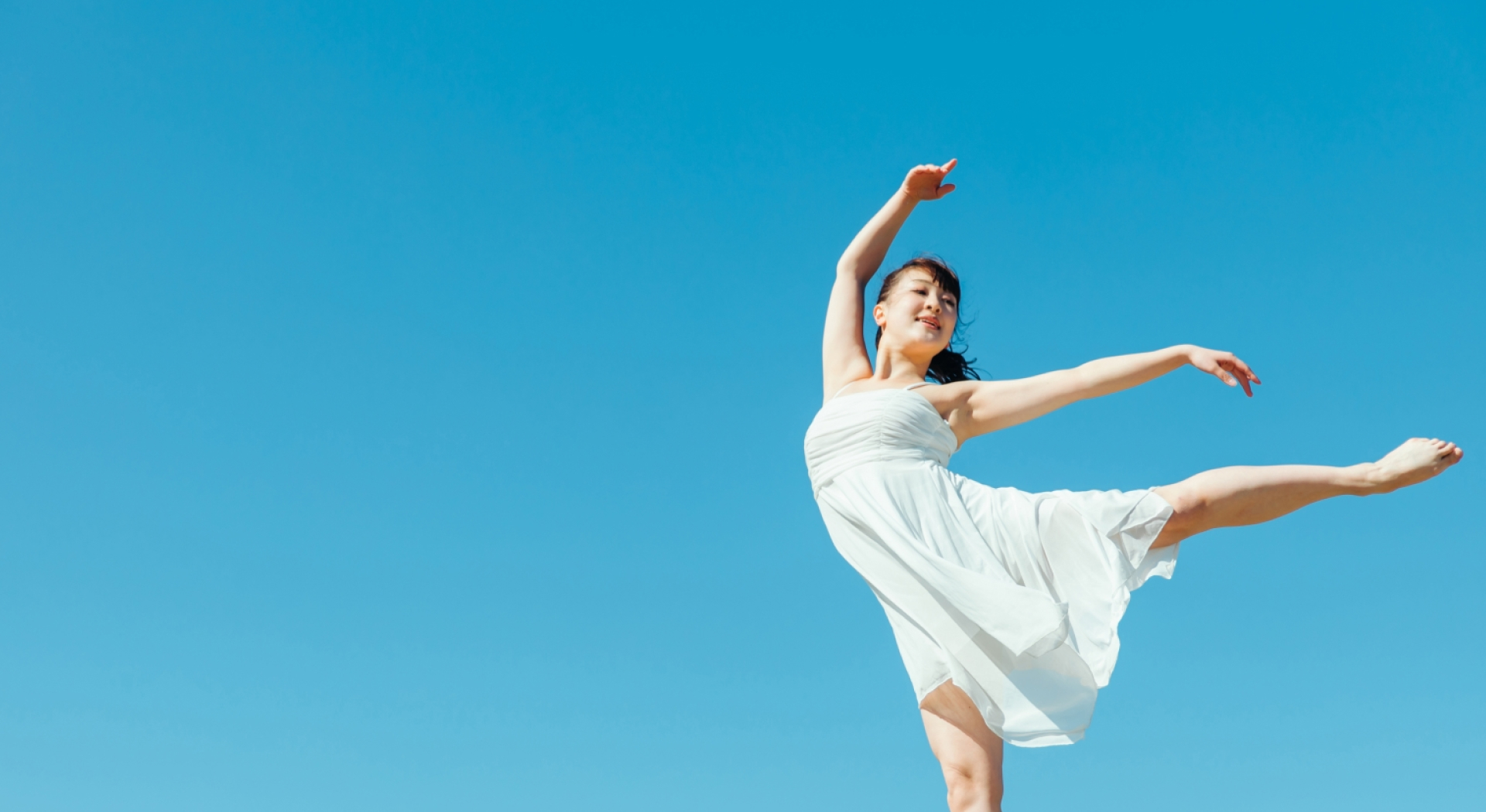 晴れた日の屋外で、白いワンピースを着た女性が片足を上げてダンスしている